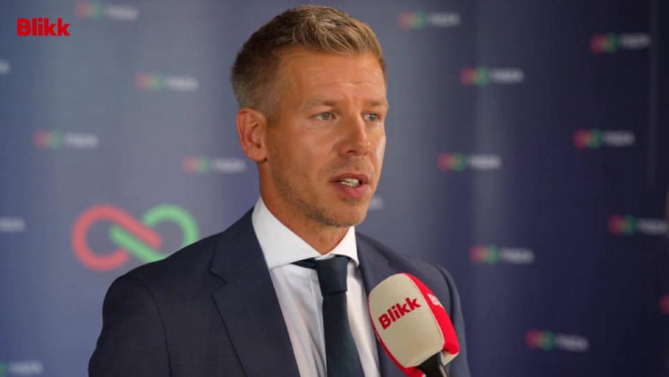 Politikus beszél egy sajtótájékoztatón, mikrofon a kezében, háttérben médiaportál logó.