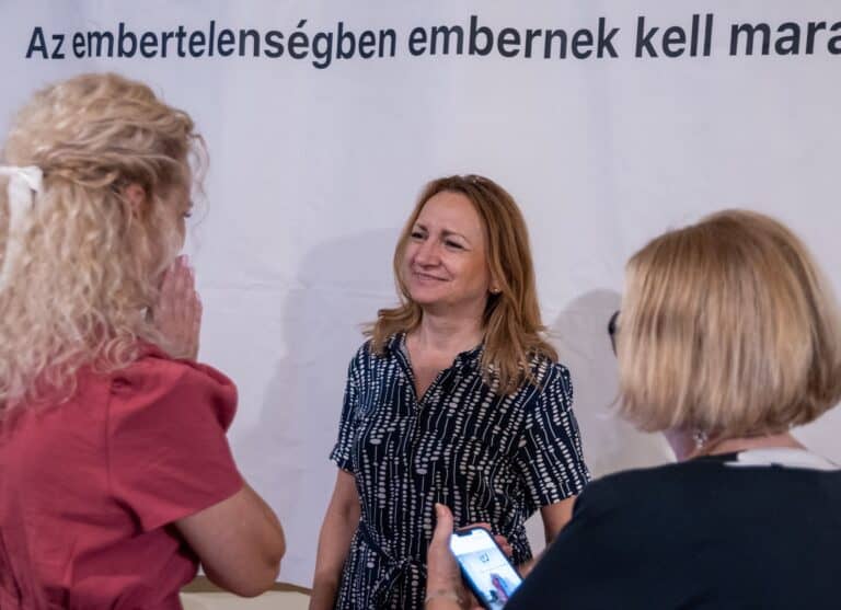 Beszélgetés három nő között, egyikük a háttérben mosolyogva, informális találkozó, baráti hangulat.