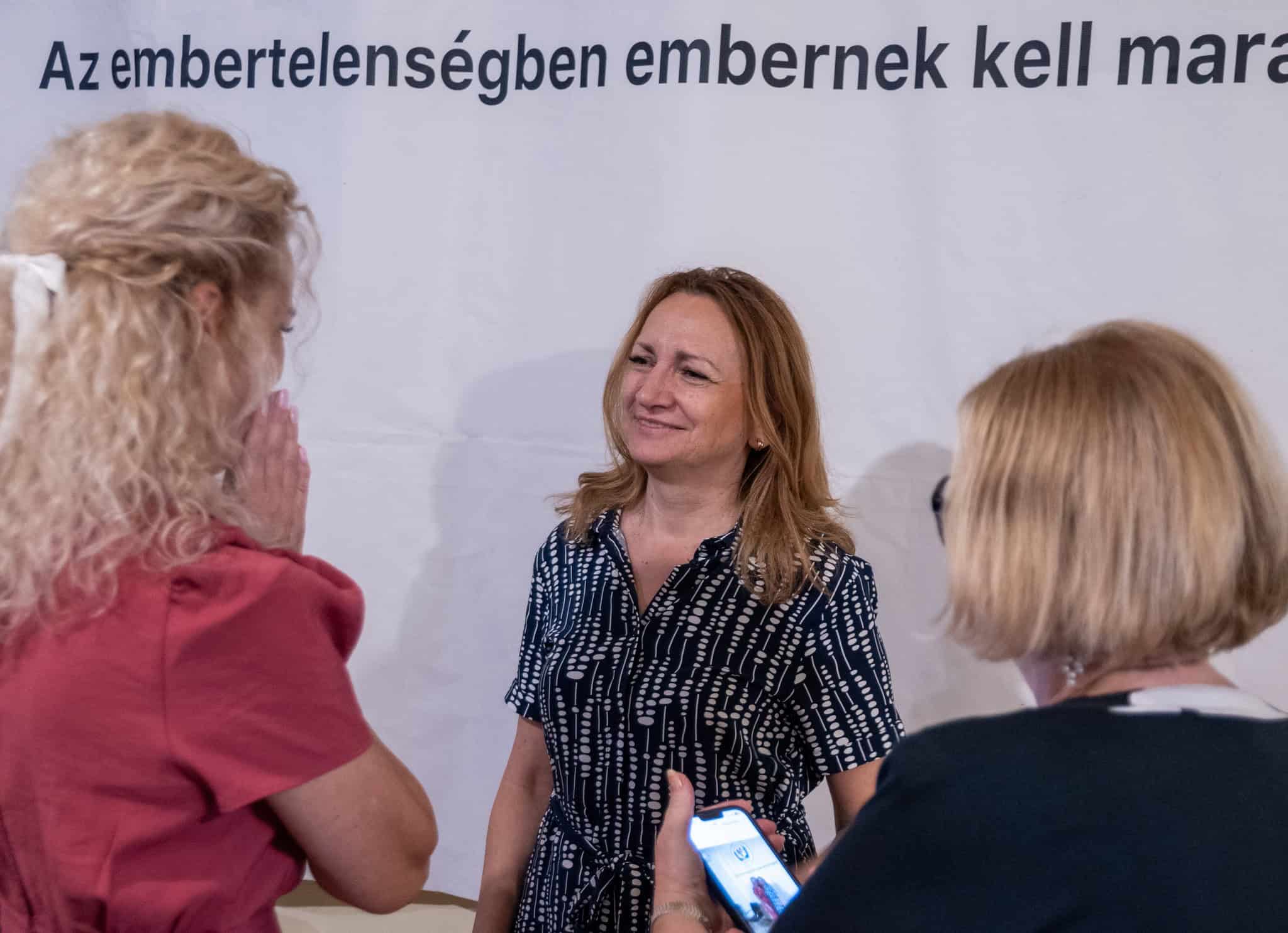 Beszélgetés három nő között, egyikük a háttérben mosolyogva, informális találkozó, baráti hangulat.