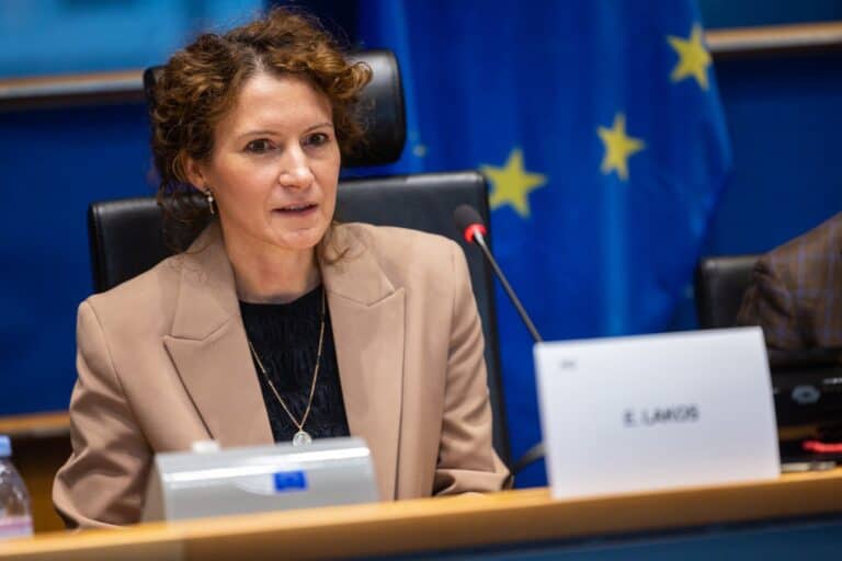 Pozícióban lévő nő európai uniós ülésen, háttérben EU zászló.