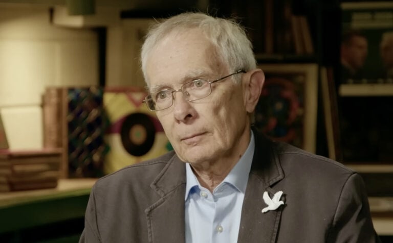 Művész portréja egy könyvespolc előtt, komoly arckifejezéssel, elegáns ruhában, diadém a zakóján.