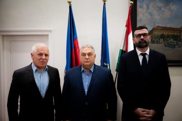 Három férfi áll egy szobában, háttérben magyar és uniós zászlók, komoly hangulat.