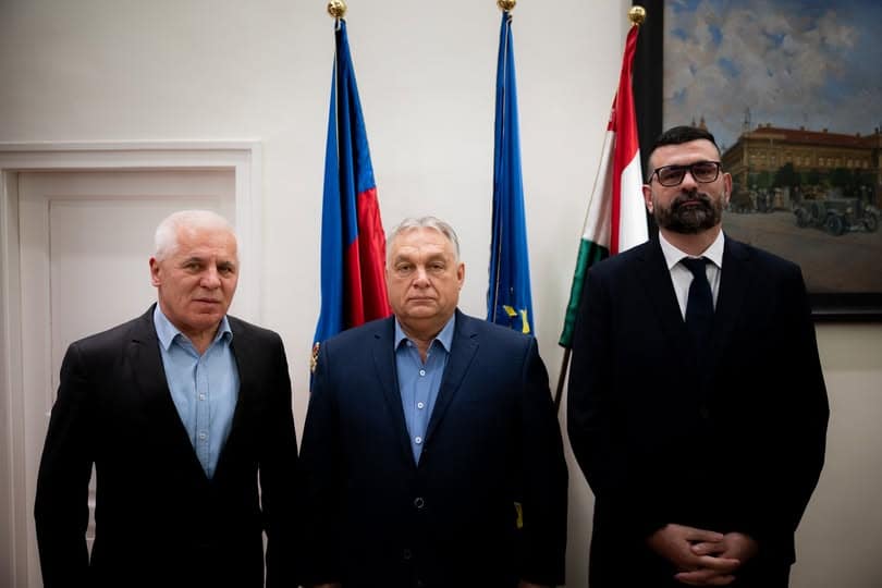 Három férfi áll egy szobában, háttérben magyar és uniós zászlók, komoly hangulat.