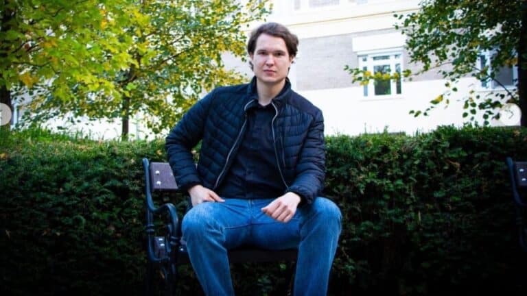 Fiatal férfi ül egy park ban, ültetvények és házak mögött.