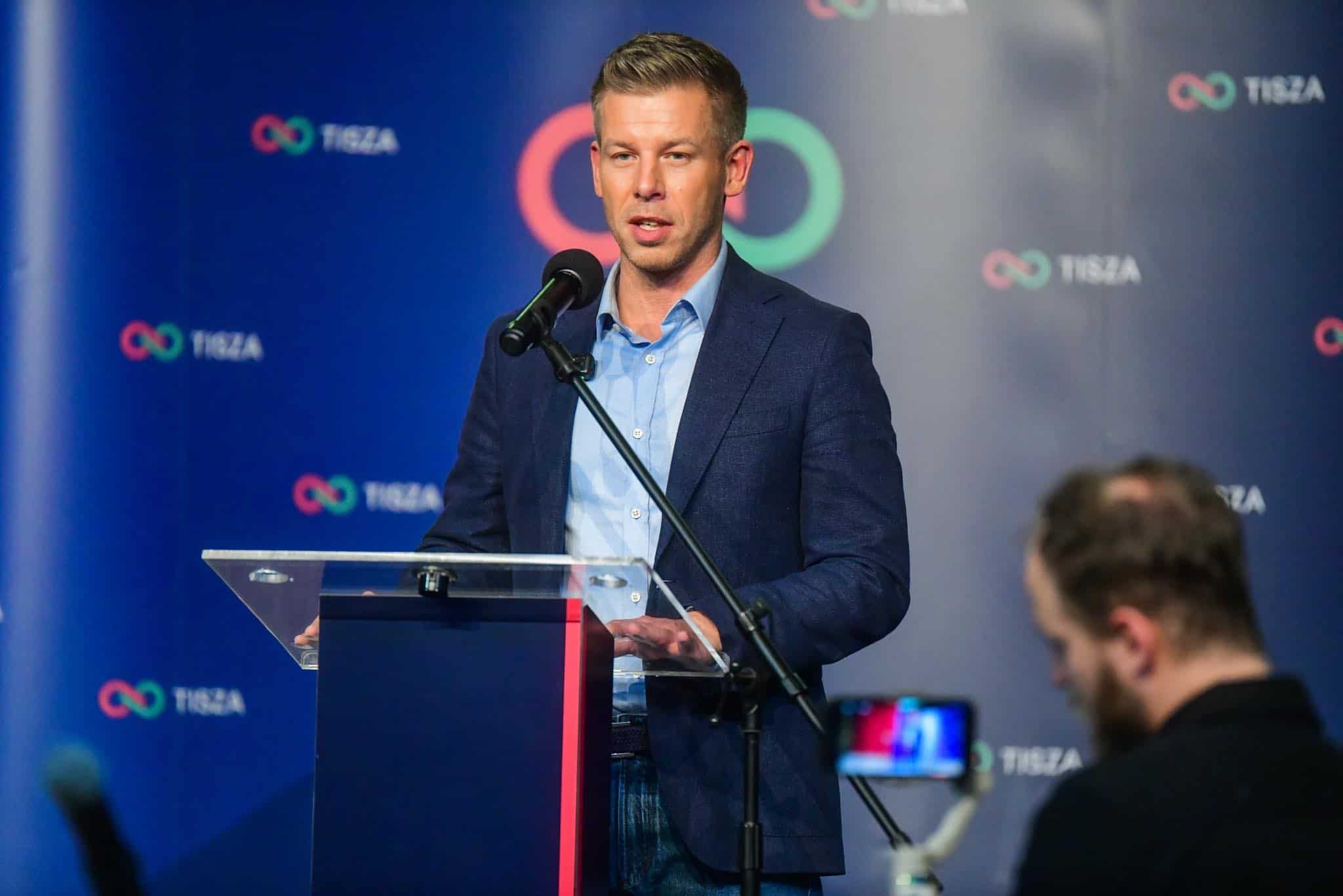 Országos vezető beszédet mond egy konferencián, mögötte a Tisza logo látható.