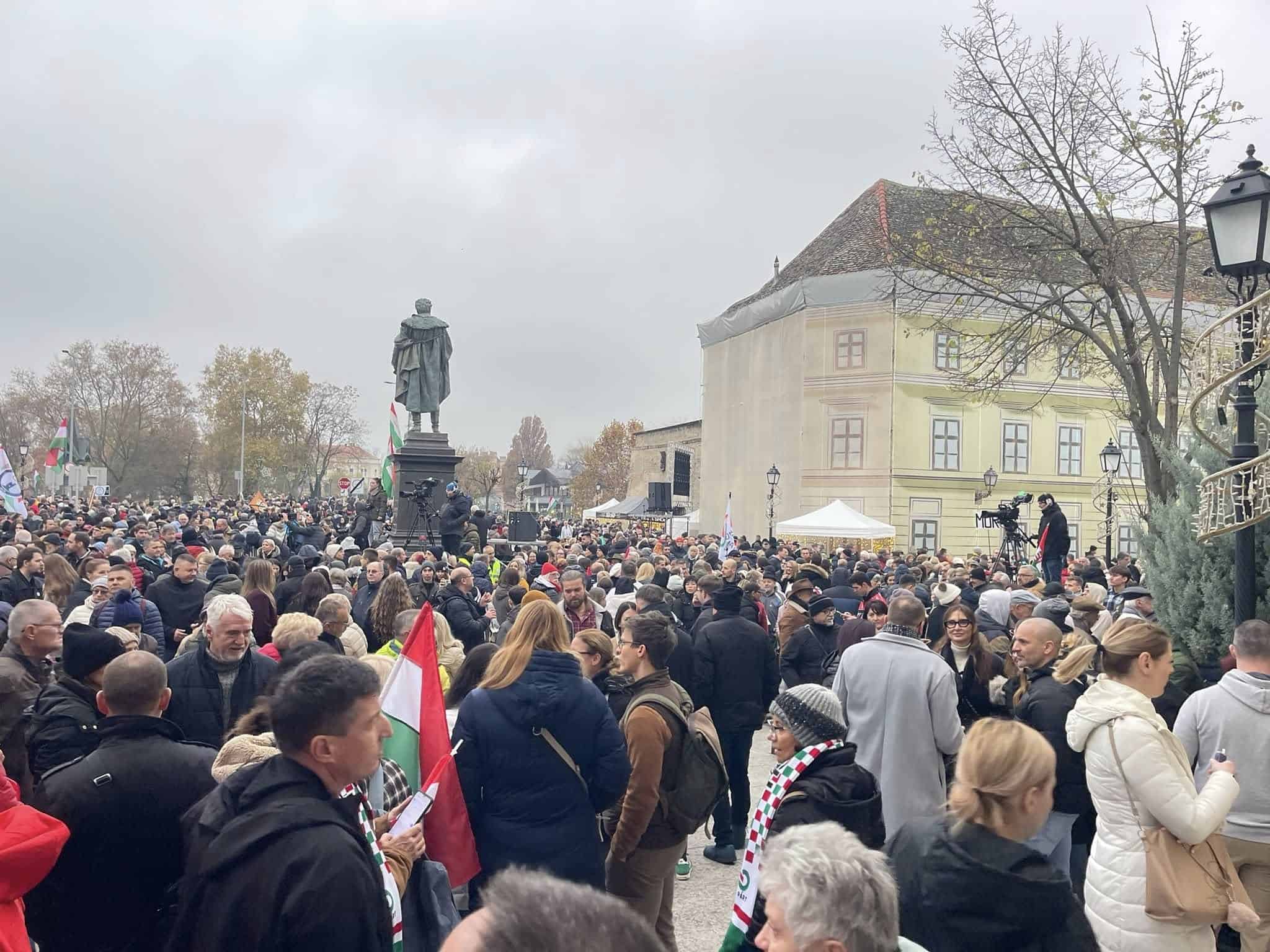 Közönség a területen, szürke ég, városi légkör, magyar zászlók, nyilvános esemény, tömeges összejövetel.
