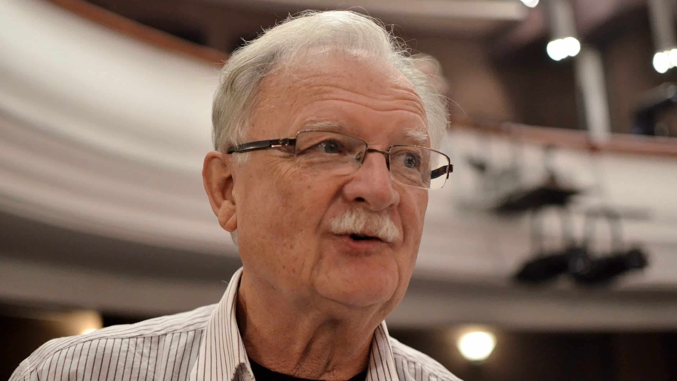 Idős férfi szemüvegben, beszélgetés közben egy rendezvényen.