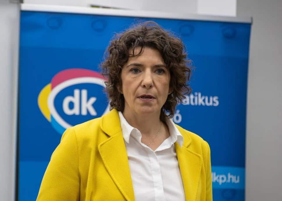 Bartók Andrea, a DK politikusa beszédet tart egy sajtótájékoztatón, háttérben a párt logójával.