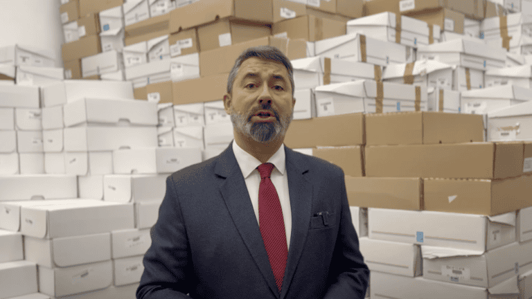 Áramszámlát tartó férfi az áruház raktárában, sok csomag és doboz a háttérben.