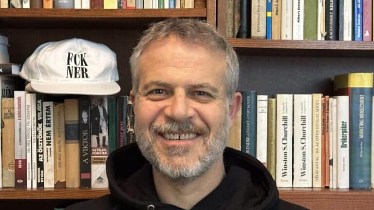 Férfi portré könyvesbolt előtt, háttérben könyvespolc, mosolygó arc, informális megjelenés.