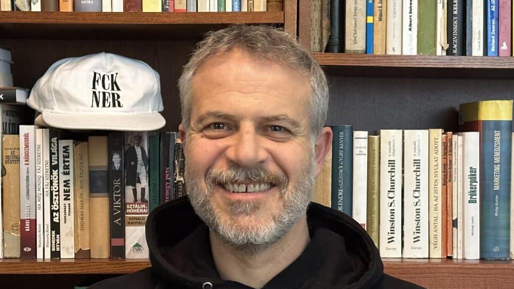 Férfi portré könyvesbolt előtt, háttérben könyvespolc, mosolygó arc, informális megjelenés.