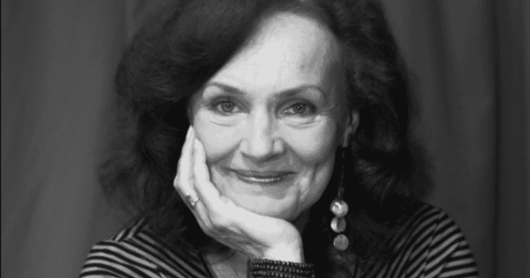 Mélyen mosolygó nőt ábrázoló fekete-fehér portré, elegáns megjelenéssel.