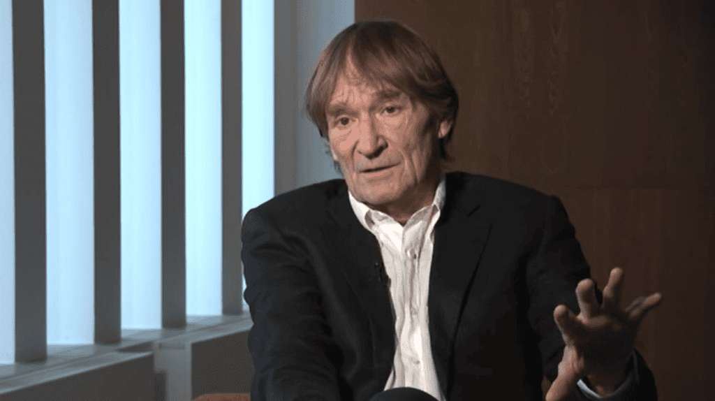 Egy idős férfi interjúban beszél, elegáns ruhában, háttérben modern ablakokkal.