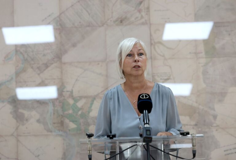 Politikus asszony beszédet tart mikrofon előtt a háttérben térkép.