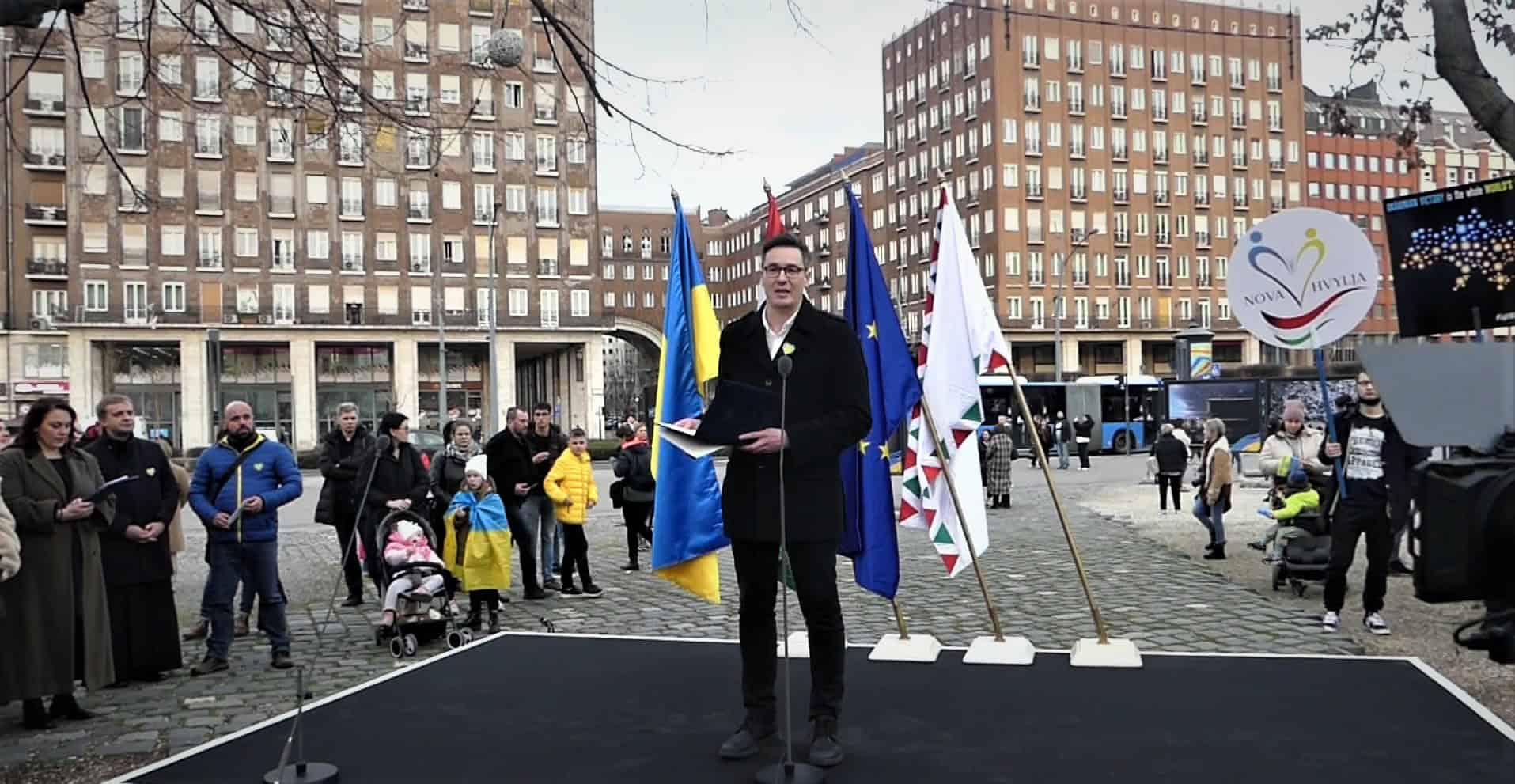 Ukrán zászló és EU zászló a háttérben, beszédet mondó férfi a nyilvános megemlékezésen, városi környezetben.