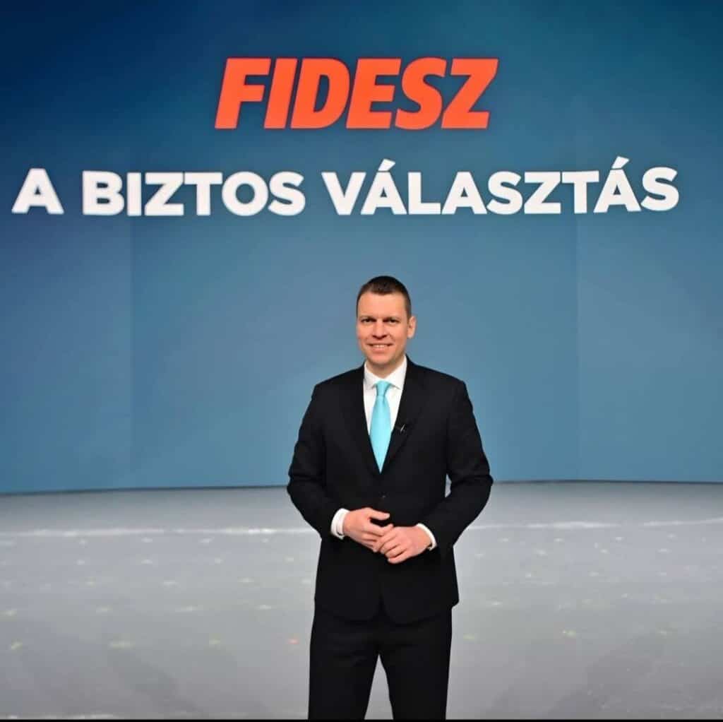 Az a képen egy férfi áll, fekete öltönyben, fehér ingben és kék nyakkendőben, háttérben a FIDESZ-es kampány szlogennel.