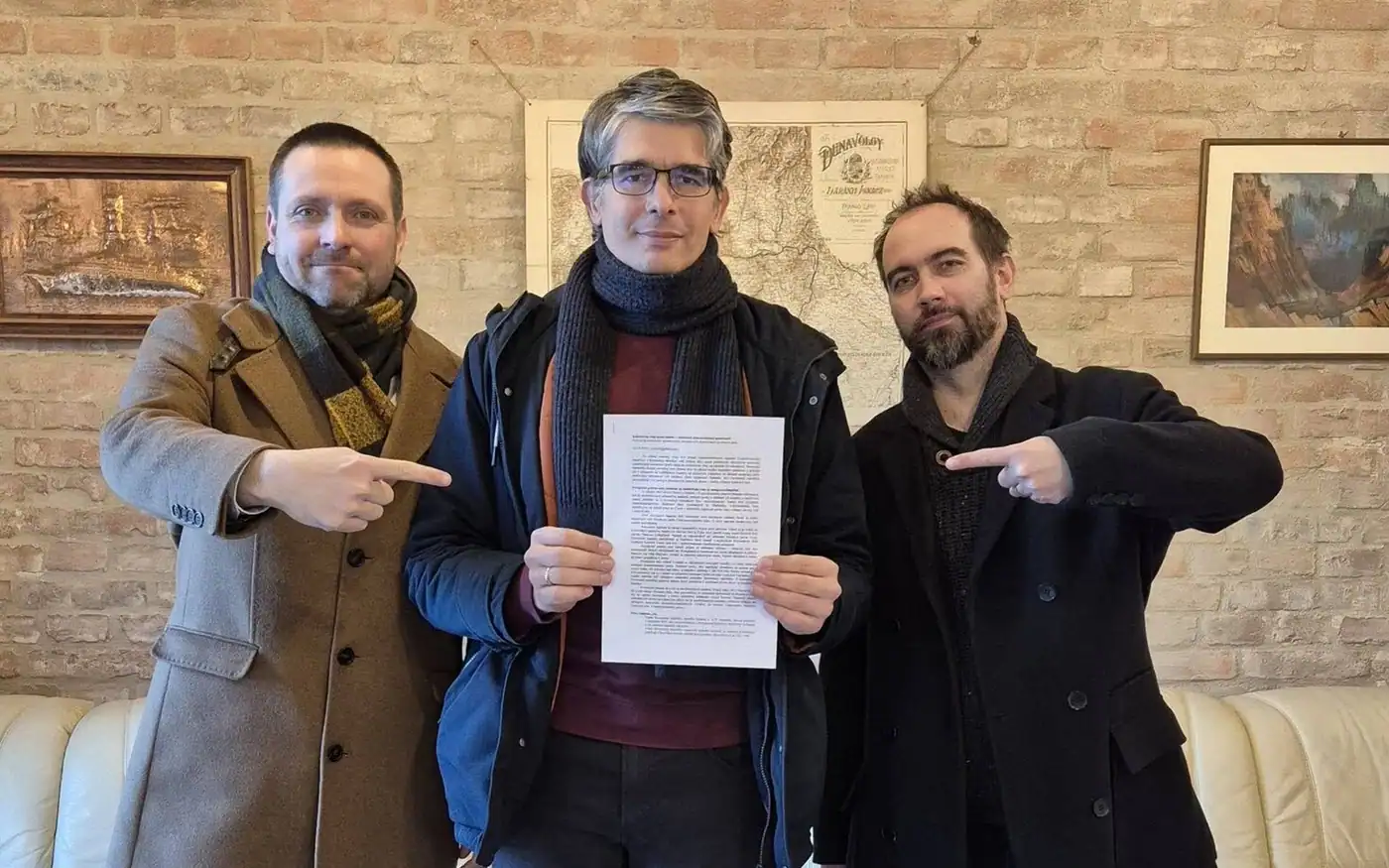 Széleskörű dokumentumot mutatnak a férfiak, akik közül a középen álló tartja a papírt.