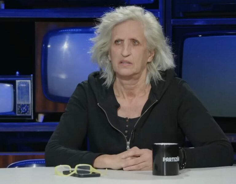 Szomorú női arc, fekete hoodie, retro tévék és kávésbögre a háttérben, komoly hangulat.