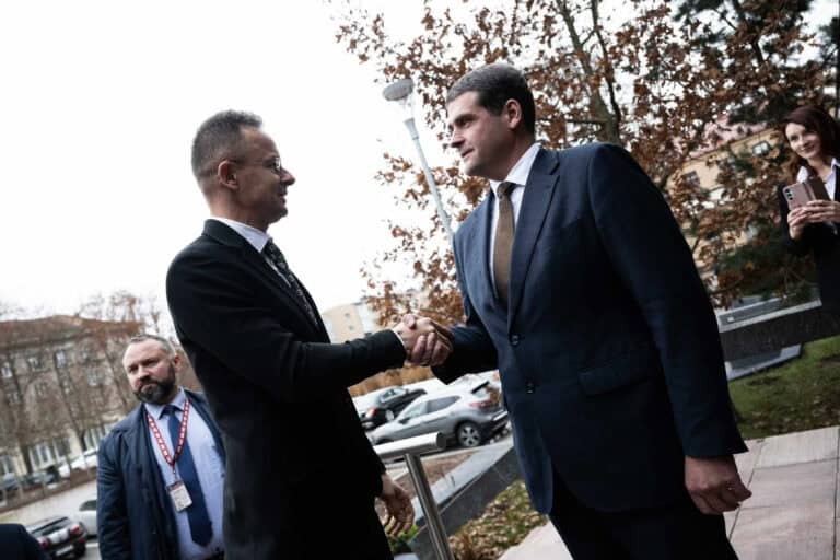 Egyeztető találkozó, két üzletember kezet fog, dokumentumok és egy távoli épület a háttérben.