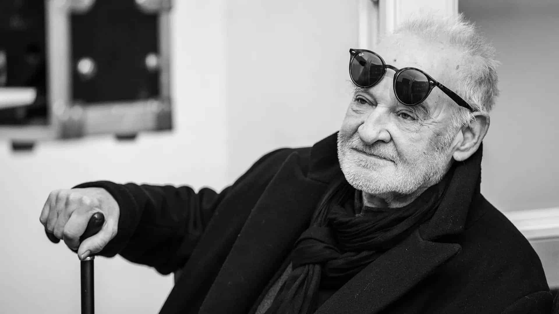 Életvidám idős férfi, napszemüveg a fején, sétáláshoz készült botot tart.