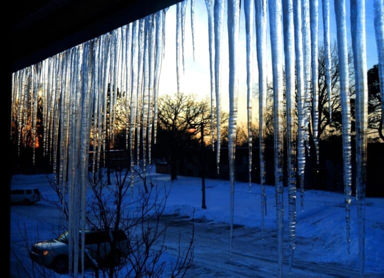 Havas jégcsapok lógnak az ablakról téli naplementében, hóval borított kert látképe.