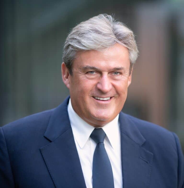 Politikus férfi elegáns öltönyben, vidám mosollyal, kék háttér előtt.