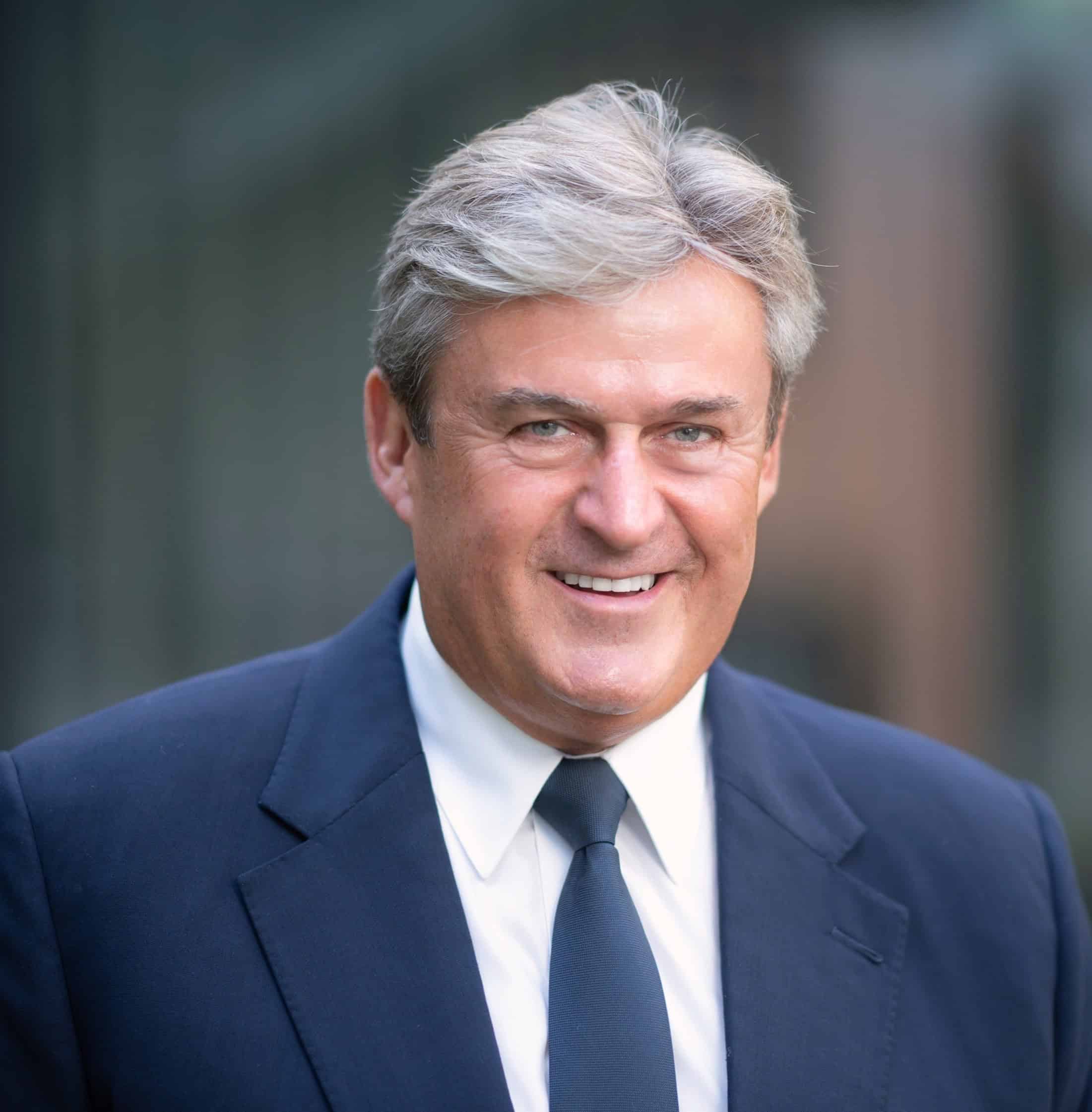 Politikus férfi elegáns öltönyben, vidám mosollyal, kék háttér előtt.