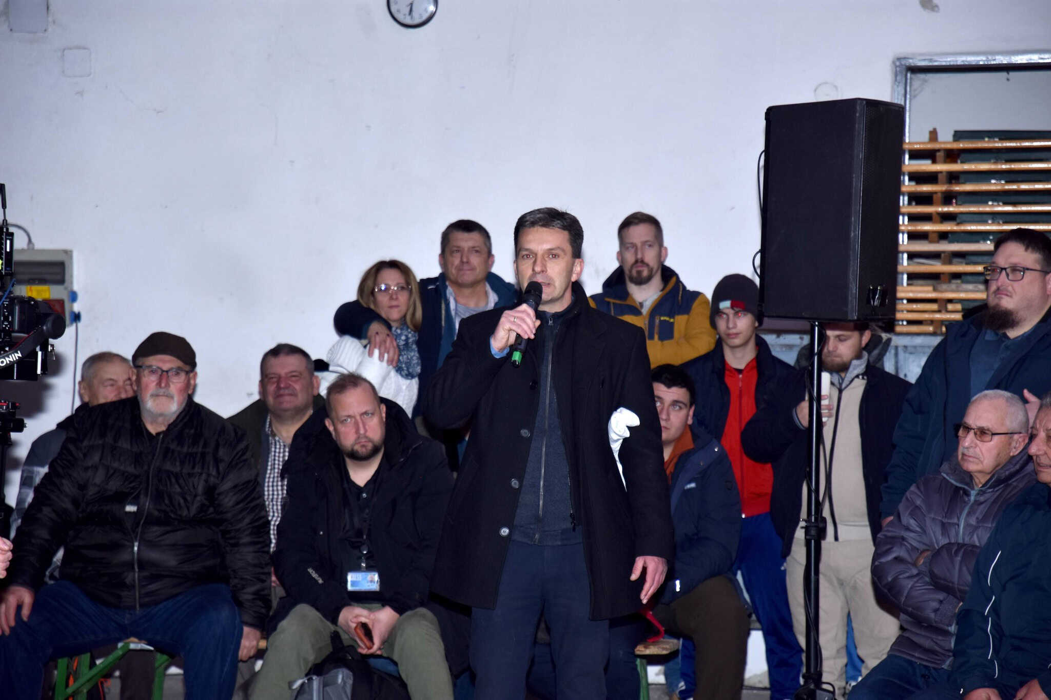 Fiatal férfi beszél egy rendezvényen, háttérben több résztvevő és hangtechnika.