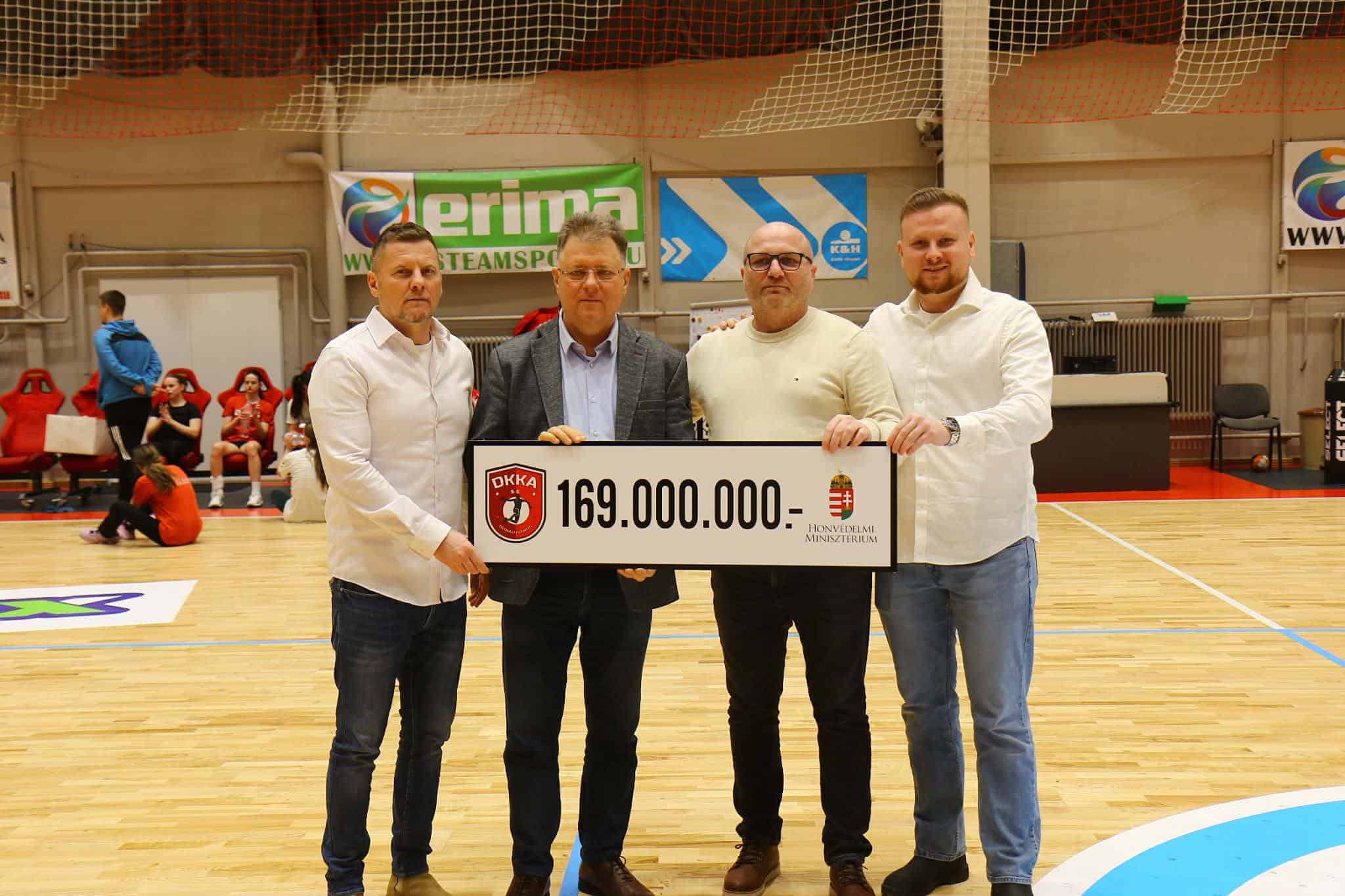 Négy férfi tart egy nagy csekket a kézilabda csapatnak a sportcsarnokban.