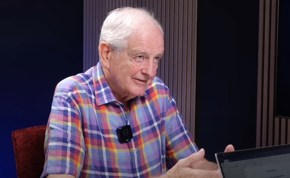 Álló férfi, színes ingben, beszélgetést folytat, háttérben modern stúdiókörnyezet.