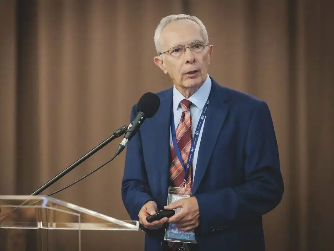 Politikus beszél egy konferencián, miközben mikrofont és jegyzeteket tart a kezében.