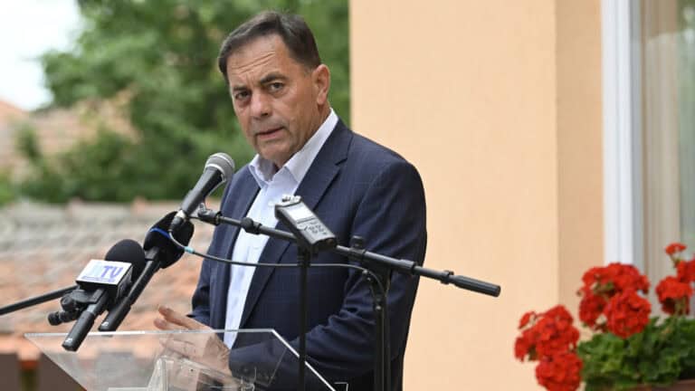 Pócs János beszél sajtótájékoztatón, háttérben virágok és épület.