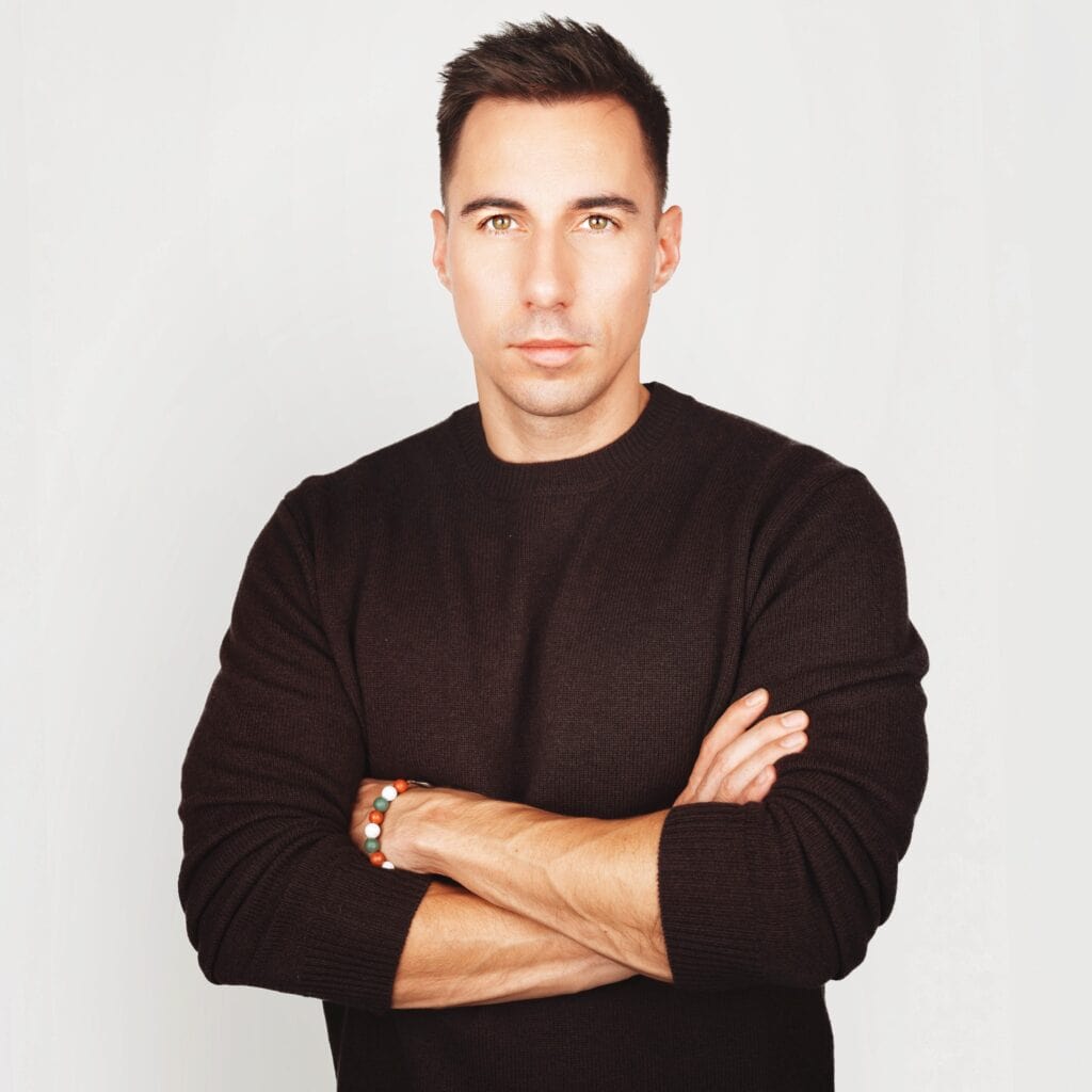 Radnai Márk fekete pulóverben, karjait keresztbe téve, komoly arckifejezéssel néz a kamerába.