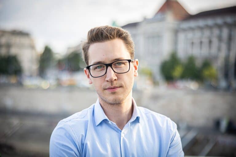 Fiatal férfi portréja, aki városban áll, háttérben épületek és folyó látható.