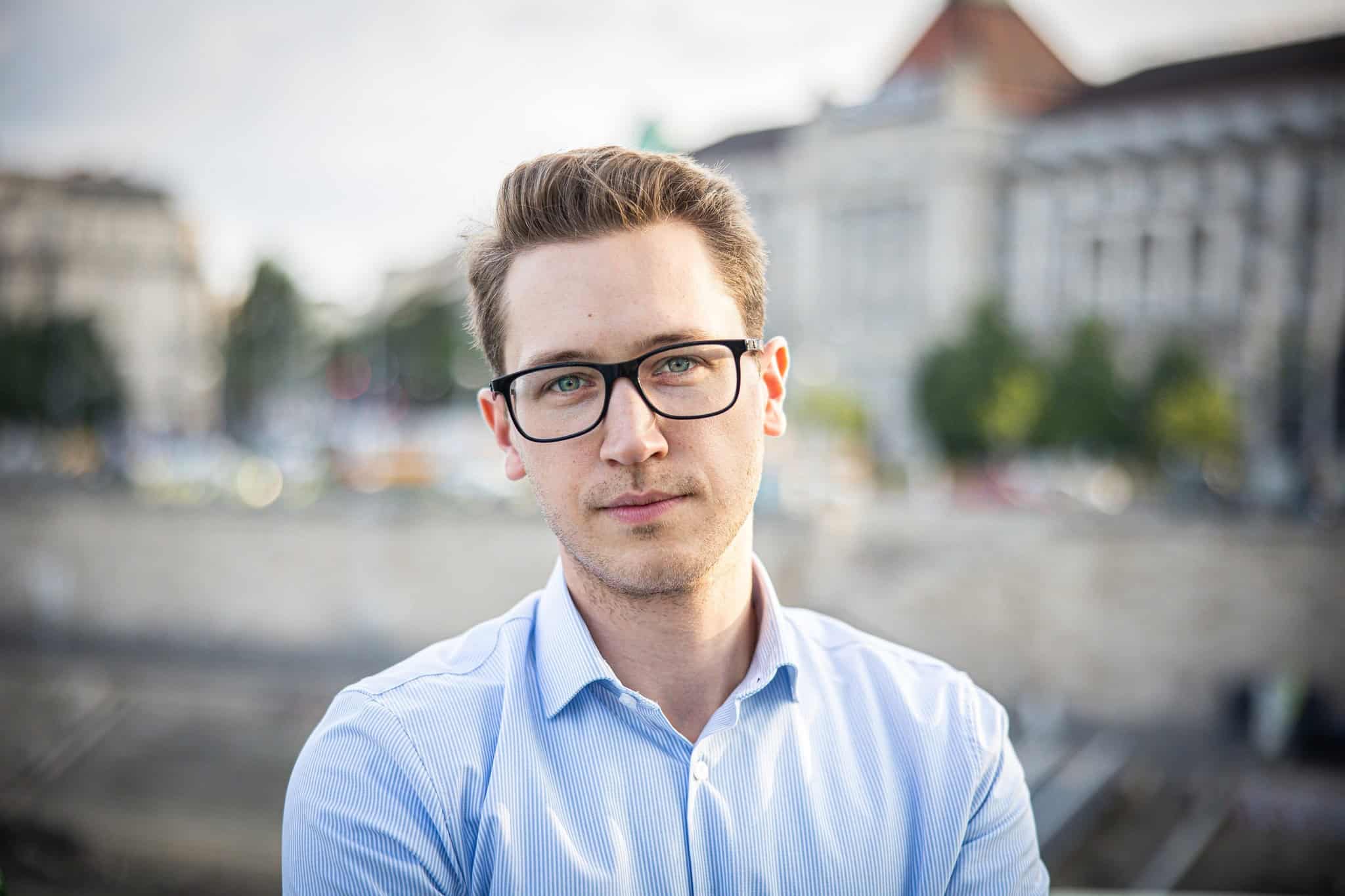 Fiatal férfi portréja, aki városban áll, háttérben épületek és folyó látható.