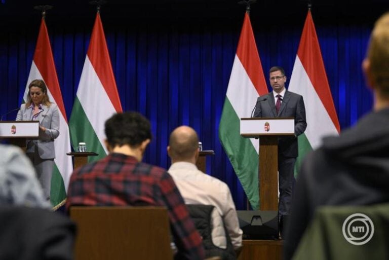 Gulyás Gergely a magyar kormány képviselője, aki sajtótájékoztatót tart, háttérben magyar zászlókkal.