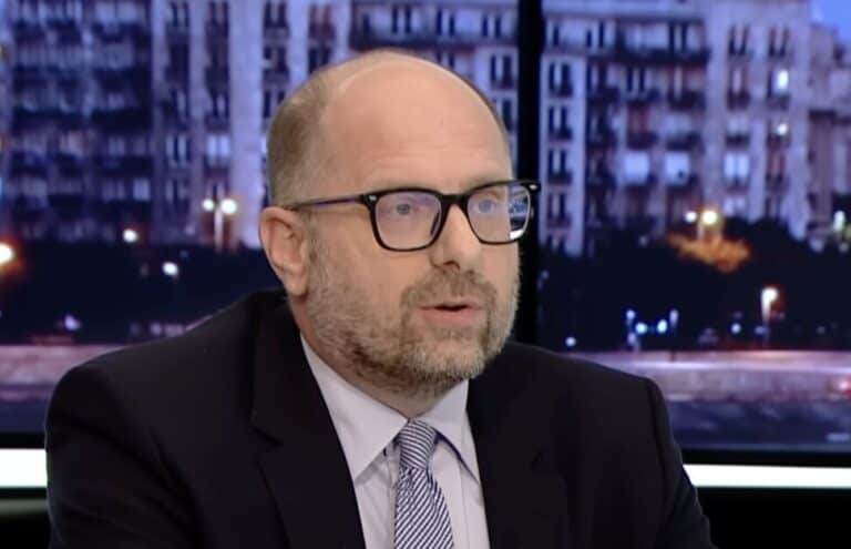 Lázár János beszél egy tévéműsorban, háttérben városi épületek.