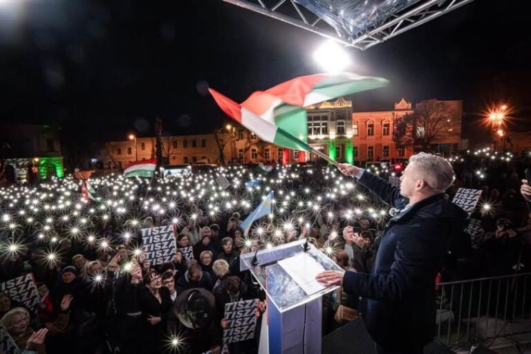 Magyar Péter a színpadon, a háttérben tömeg tüntet és magyar zászlók lobognak.