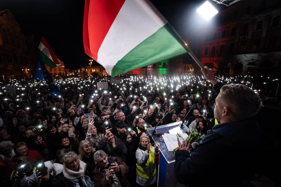 Magyar Péter a színpadon, hatalmas tömeg előtt, akik a Széchenyi téren hallgatják beszédét.