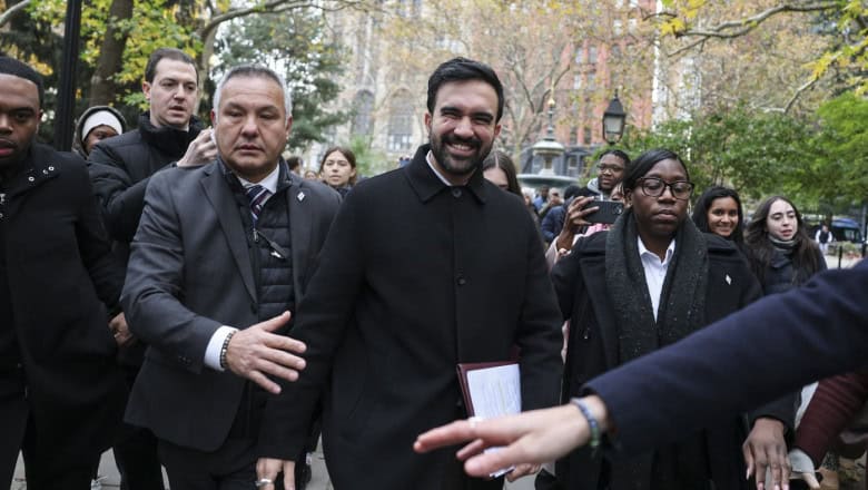 Embercsoport a robbanás helyszínén, nyomozók és médiások a közelben.