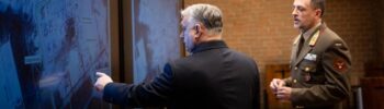 Két férfi elemzi a műholdfelvételeket egy kijelzőn, a háttérben téglafal.