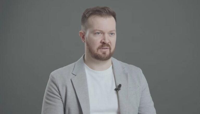 Férfi beszél egy videóinterjúban, szürke háttér előtt.