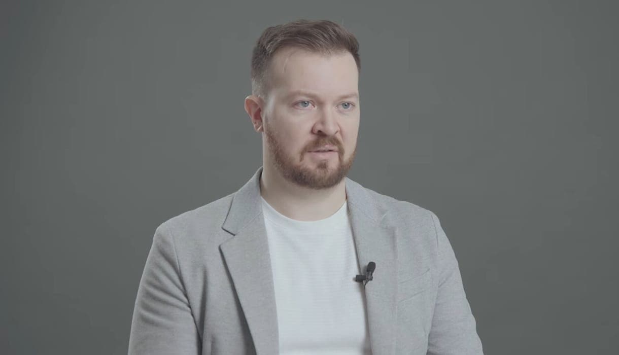 Férfi beszél egy videóinterjúban, szürke háttér előtt.