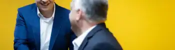 Az egyik politikus mosolyogva kezet fog egy másik személyével egy sárga fal előtt.