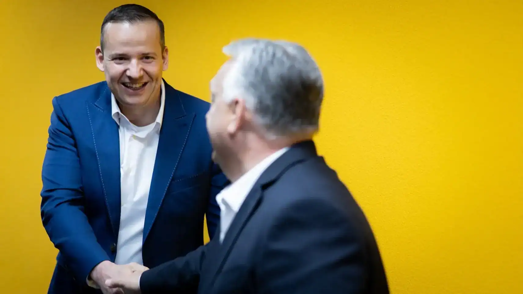 Az egyik politikus mosolyogva kezet fog egy másik személyével egy sárga fal előtt.