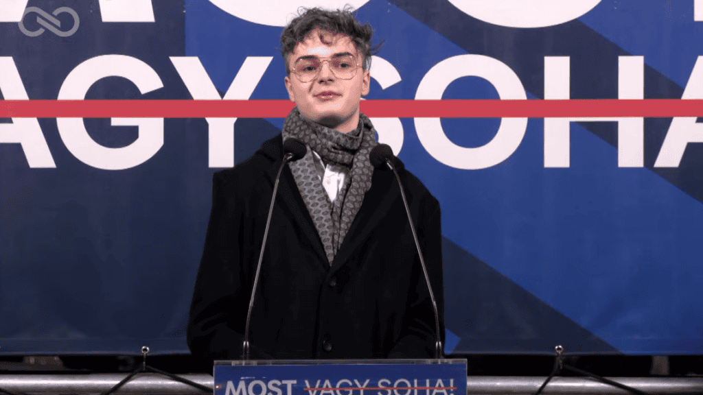 Fiatal férfi beszél egy konferencián, mikrofon előtt állva.