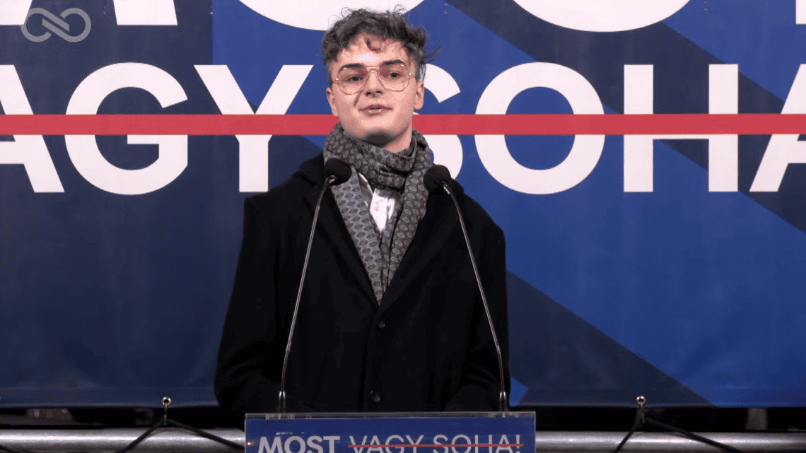 Fiatal férfi beszél egy konferencián, mikrofon előtt állva.