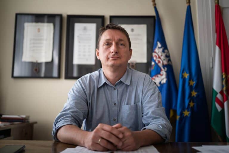 Az asztalnál ülő férfi portréja, háttérben magyar és európai uniós zászlók és oklevelek.