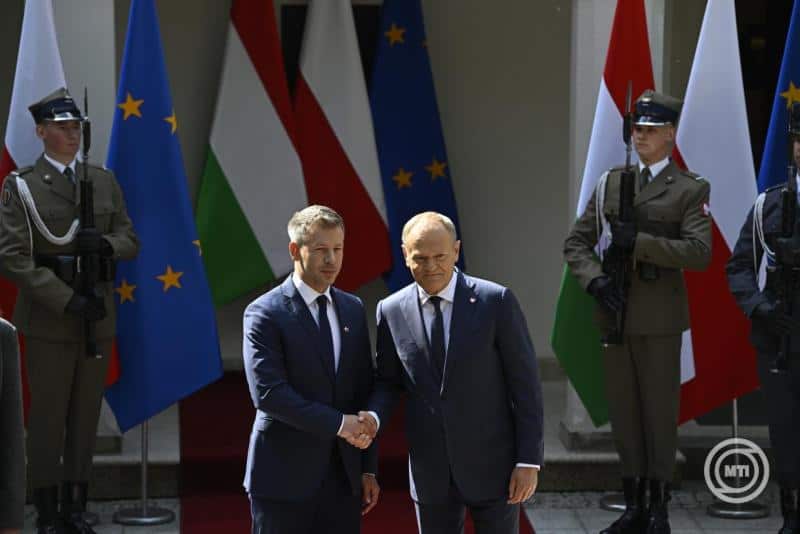 Magyar és osztrák vezetők kezet fogva, nemzetközi zászlók előtt.