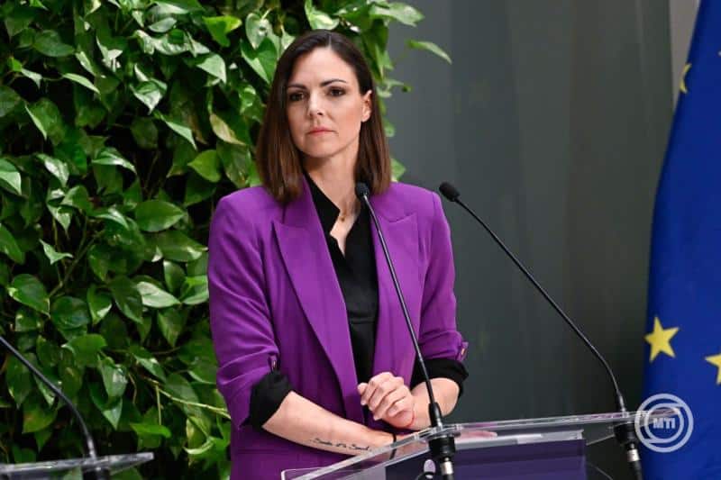 Női politikus beszél egy sajtótájékoztatón, zöld növények és EU zászló a háttérben.